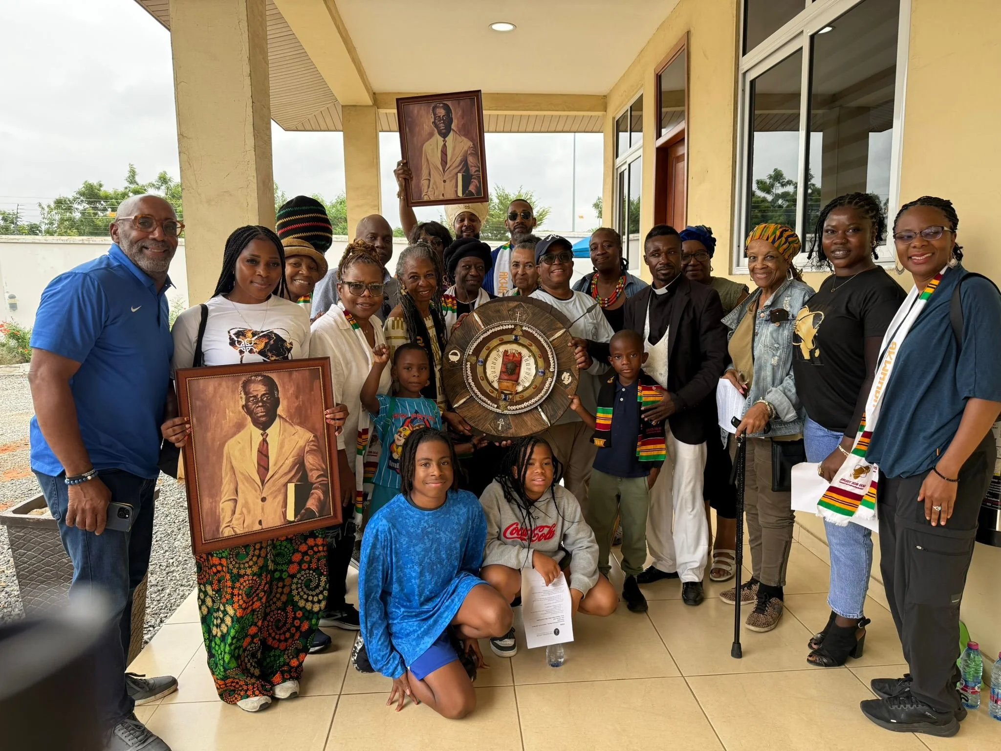Group of people posing for a photo, some holding portraits and a shield, celebrating an event related to African heritage or history. They are wearing colorful scarves and traditional attire, standing on a covered porch with a building behind them.
