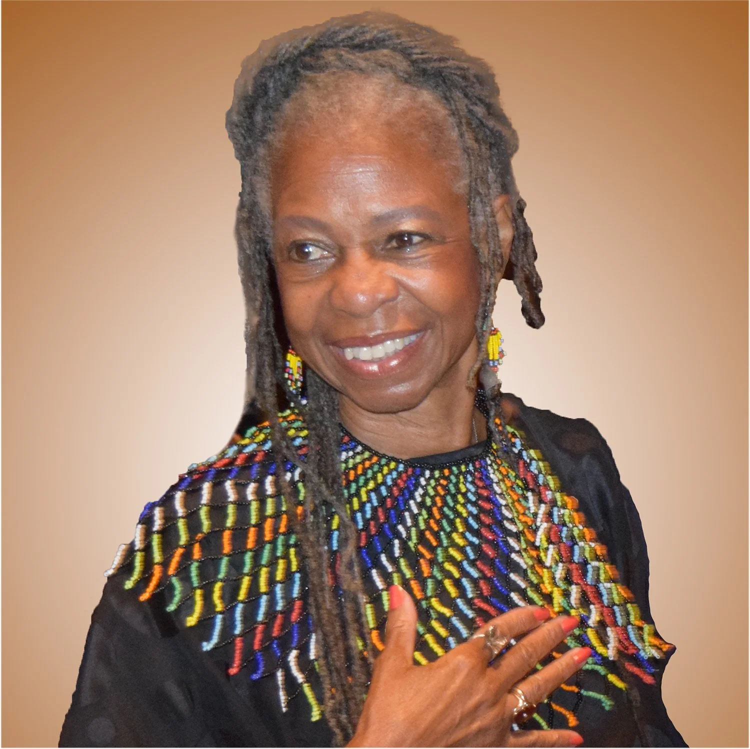 A smiling African American woman with gray dreadlocks, wearing colorful beaded jewelry and a black top, holding one hand over her chest against a neutral background.