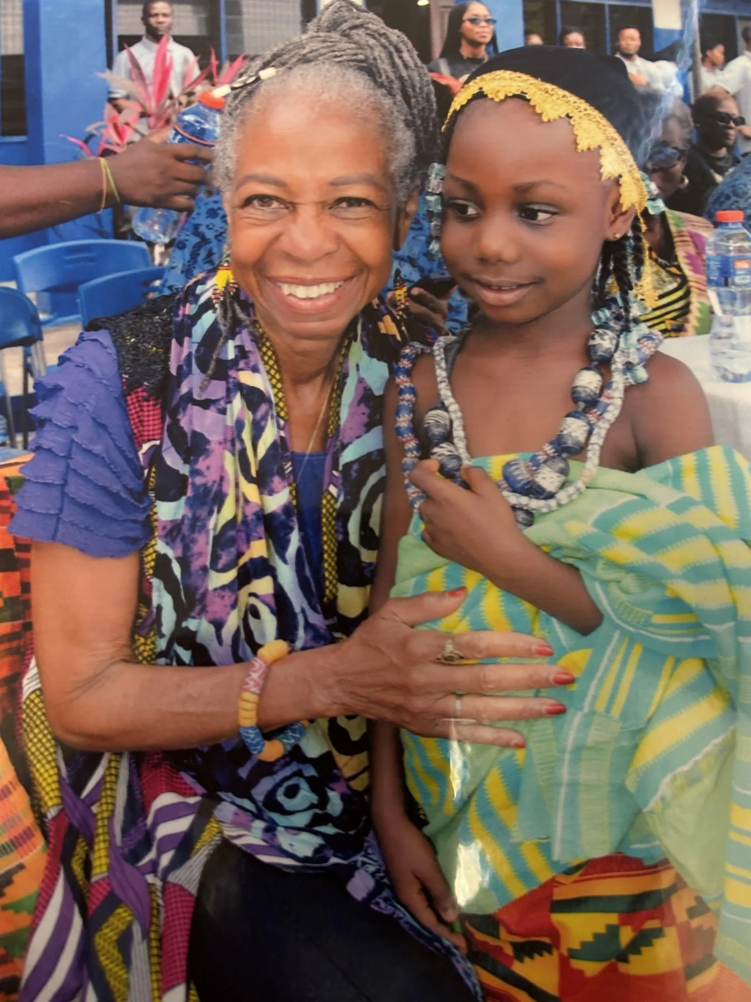 An elderly woman and a young girl wearing traditional African clothing and jewelry, sitting at a table during a celebration or cultural event.