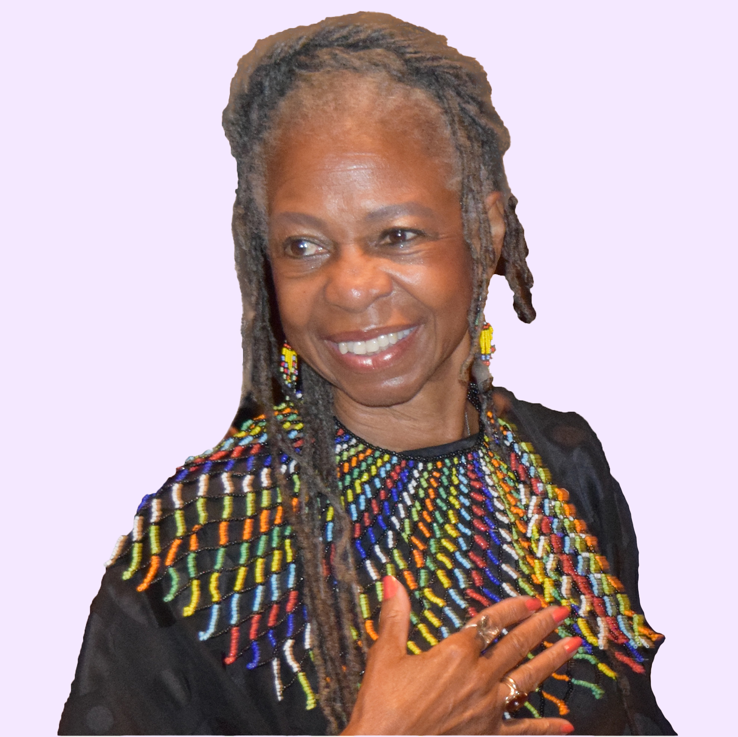 A woman with shoulder-length gray dreadlocks, smiling, wearing colorful beaded jewelry including a necklace and earrings, holding her hand to her chest, against a light purple background.