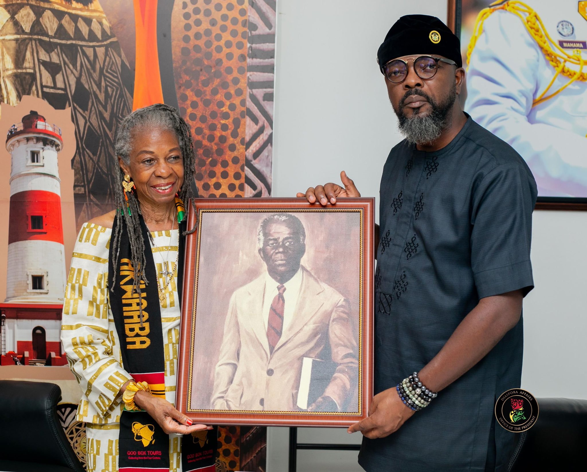 Two people holding a framed portrait of a man. The woman on the left is wearing a patterned white and yellow dress with African-inspired accessories and a black sash that says 'AFRICA.' The man on the right is dressed in a black shirt with embroidered details, glasses, and beads on his wrist and head. They are indoors with tribal art and a lighthouse mural in the background.
