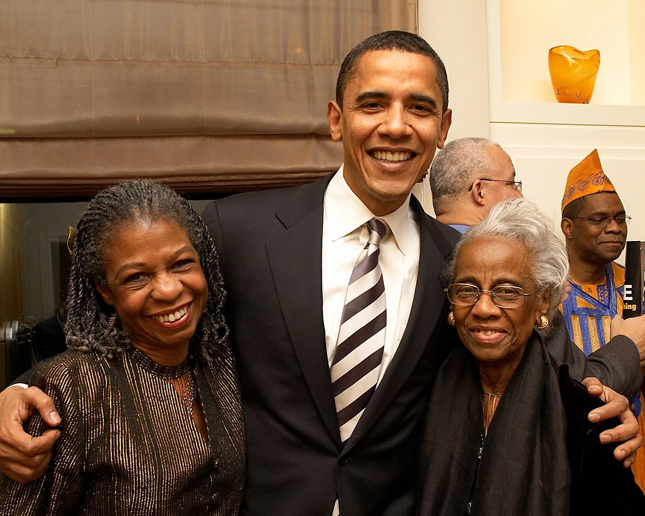 A group of four people smiling and posing for a photo indoors. The man in the center is wearing a black suit with a striped tie, and the women on either side are dressed in elegant clothing. In the background, two men are visible, one with glasses an