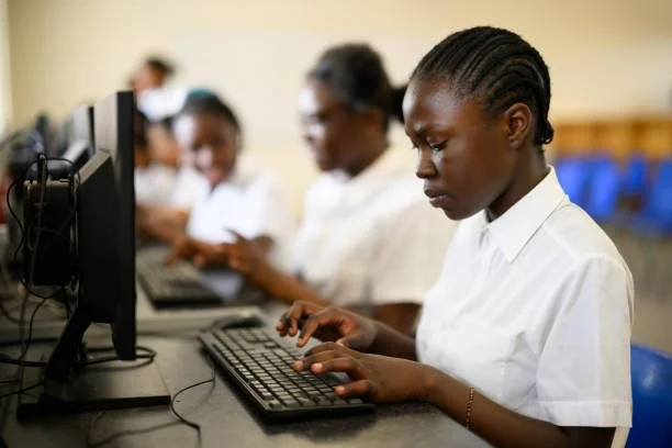 Young girl in a white shirt working on a desktop computer in a classroom with other students.