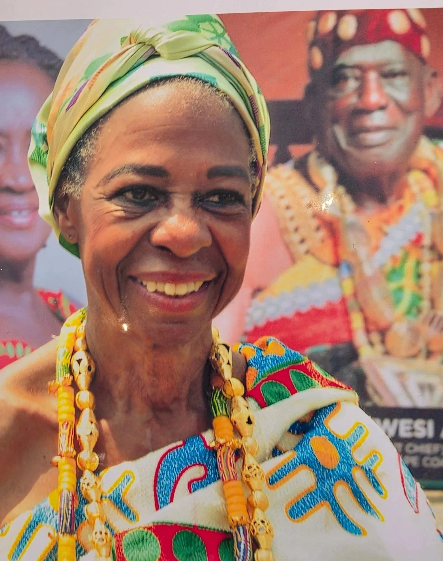 A woman wearing a colorful patterned headwrap, beaded necklace, and traditional attire, smiling in front of two other people dressed similarly and vibrant background.