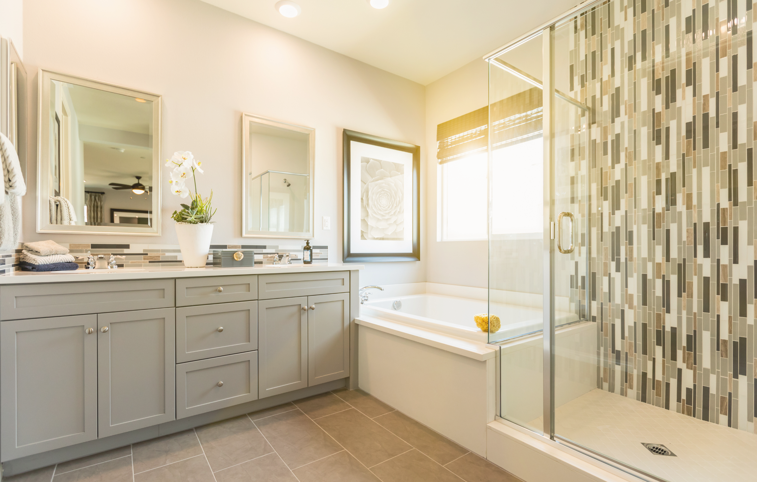 Bright bathroom with double vanity, large mirror, potted plant, framed art, bathtub by window, and walk-in shower with colorful tile wall.