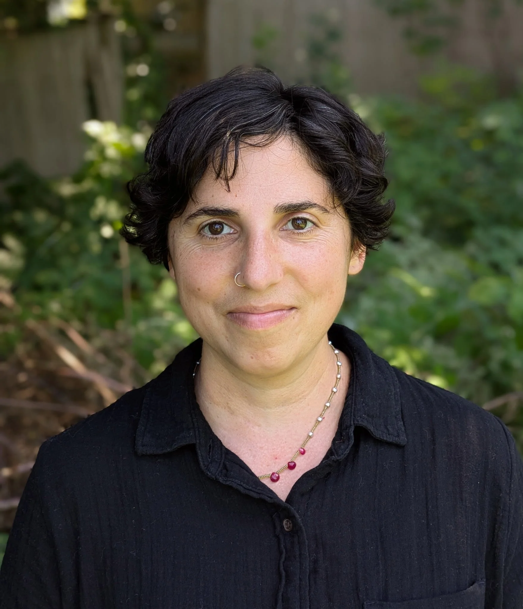 A woman with short dark hair, wearing a black shirt, a necklace, and a small nose ring, standing outdoors with greenery in the background.