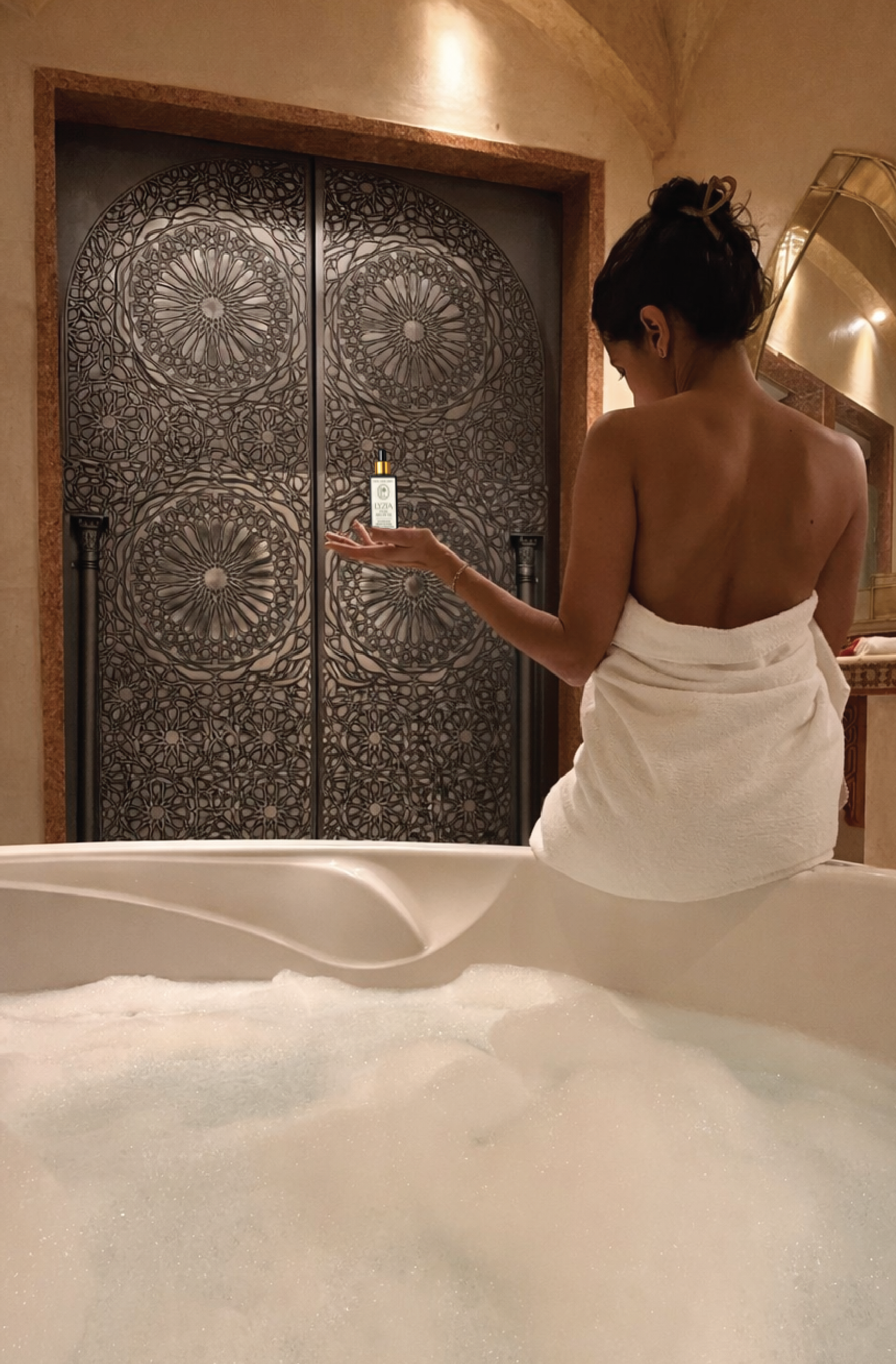 A woman sitting at the edge of a bubble-filled bathtub, wrapped in a white towel, holding a small bottle while looking at an ornate decorative door in a luxurious bathroom.