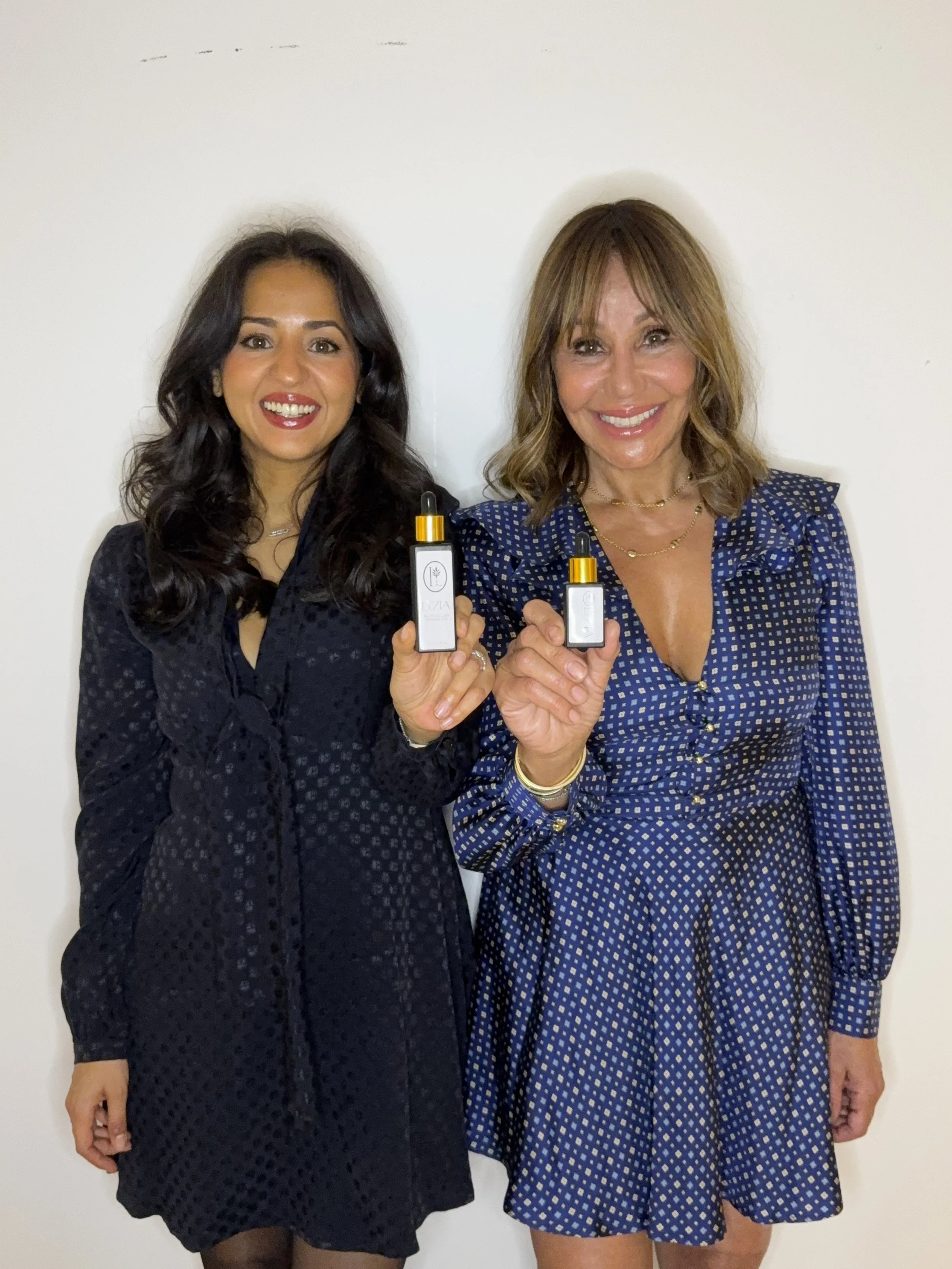 Two women smiling and holding skincare bottles, standing against a plain white wall.