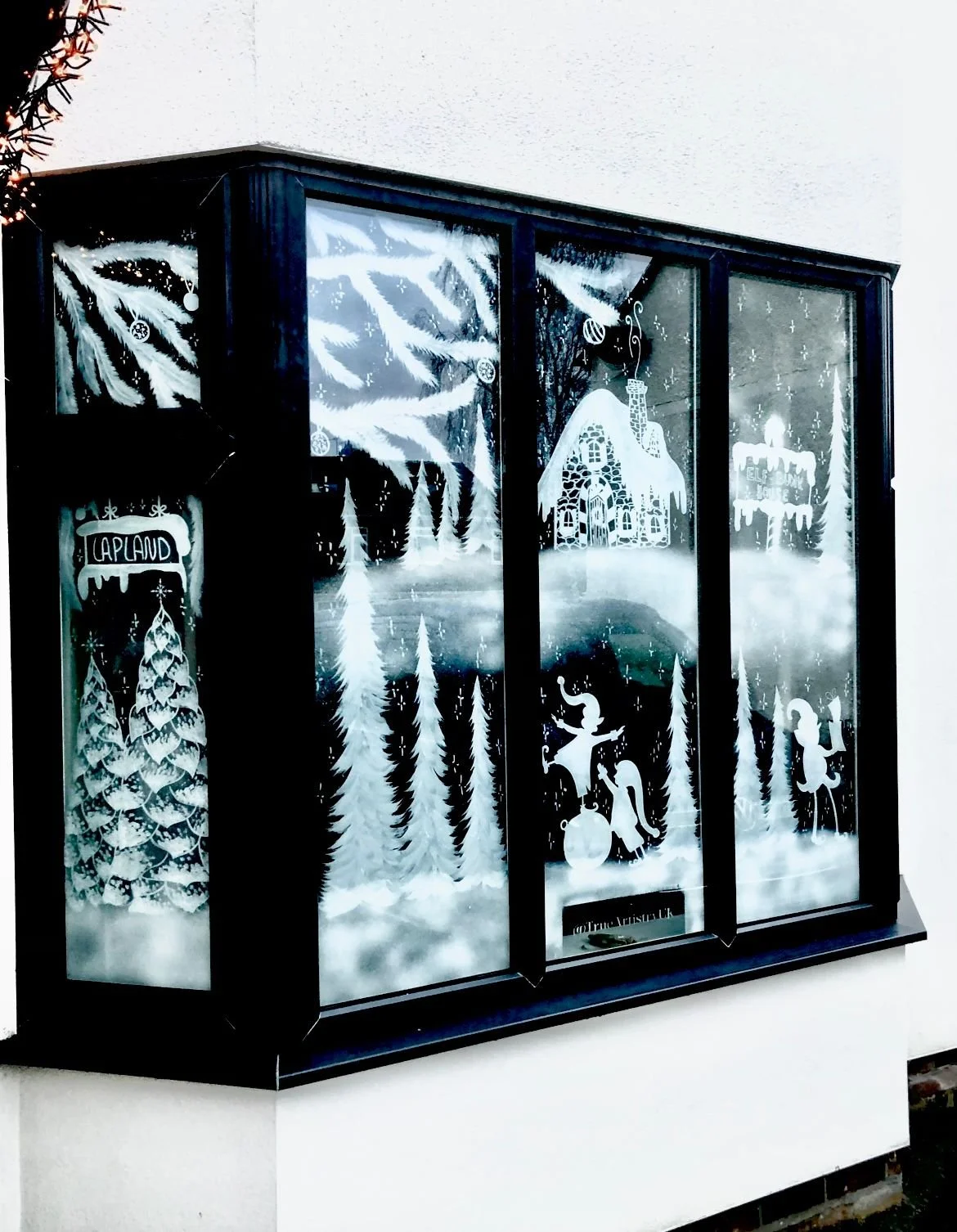 Decorative window with white Christmas-themed designs including trees, a gingerbread house, a reindeer, a girl with a lantern, and a sign reading "Caplaud," all in a winter scene.