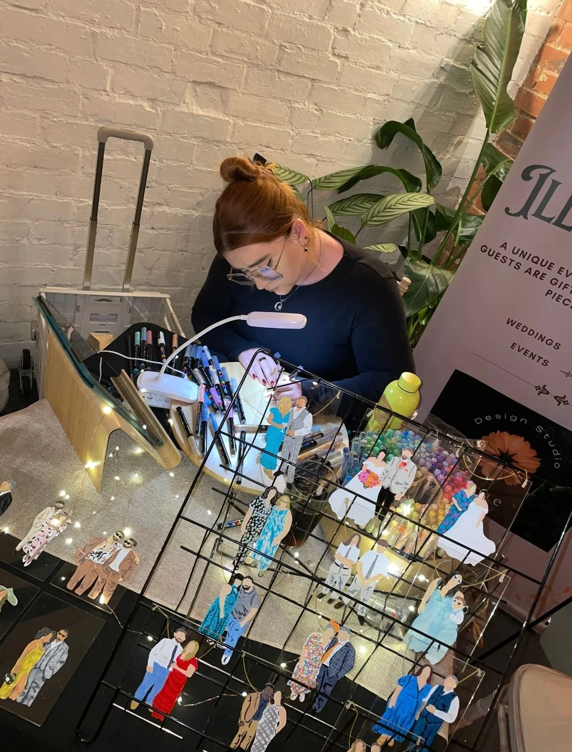 A woman with glasses and red hair working at a craft table with various art supplies, including markers and decorative cutouts of people, in a room with a white brick wall, large green plants, and a sign in the background.