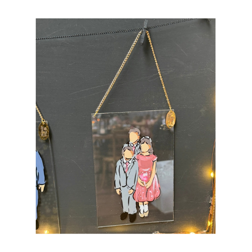 A decorative wedding guest illustration of three people, two adults and one child, hanging from a gold chain on a black wall, illuminated by soft yellow lights.