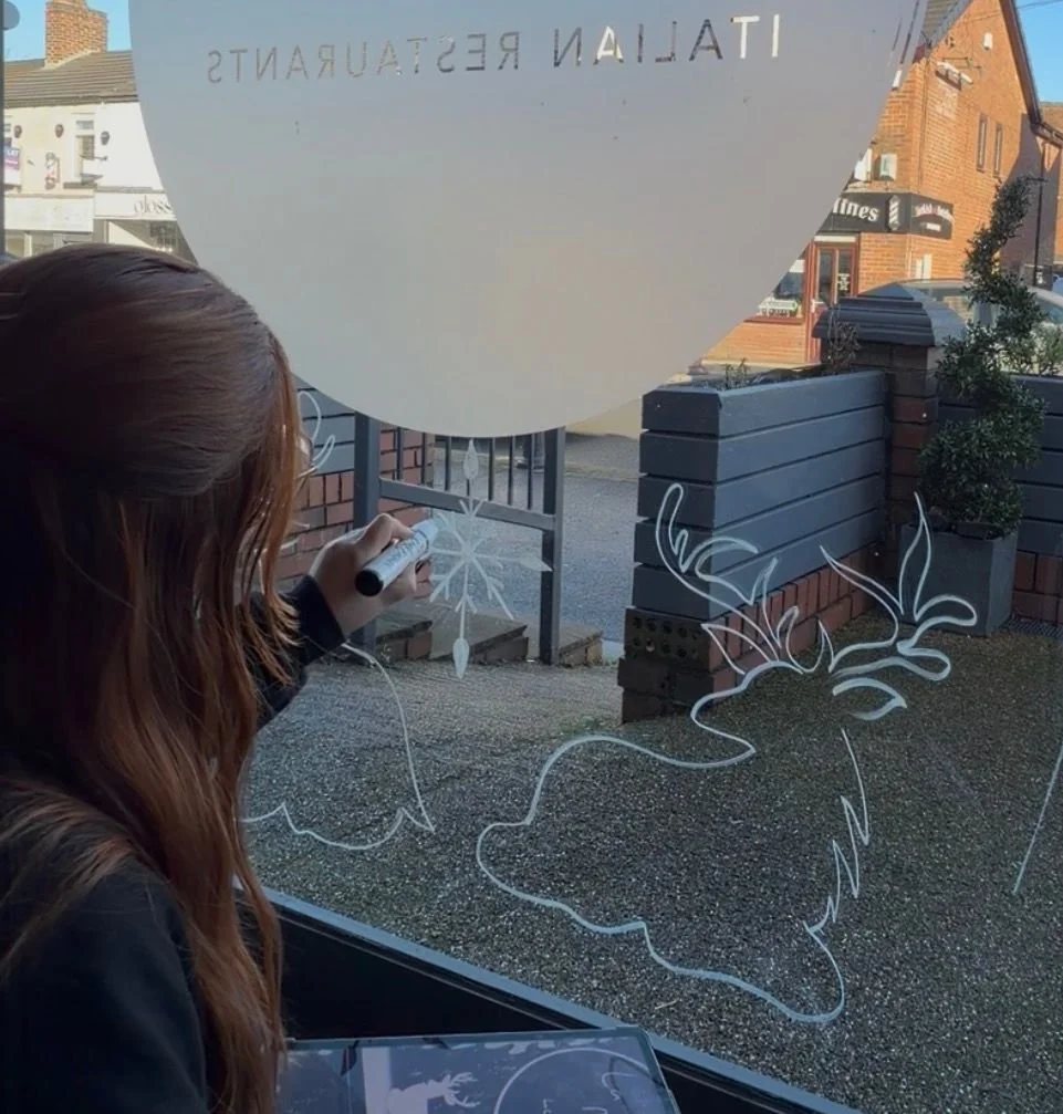 Person drawing holiday-themed white chalk designs on a window, including a reindeer with antlers and a snowflake.