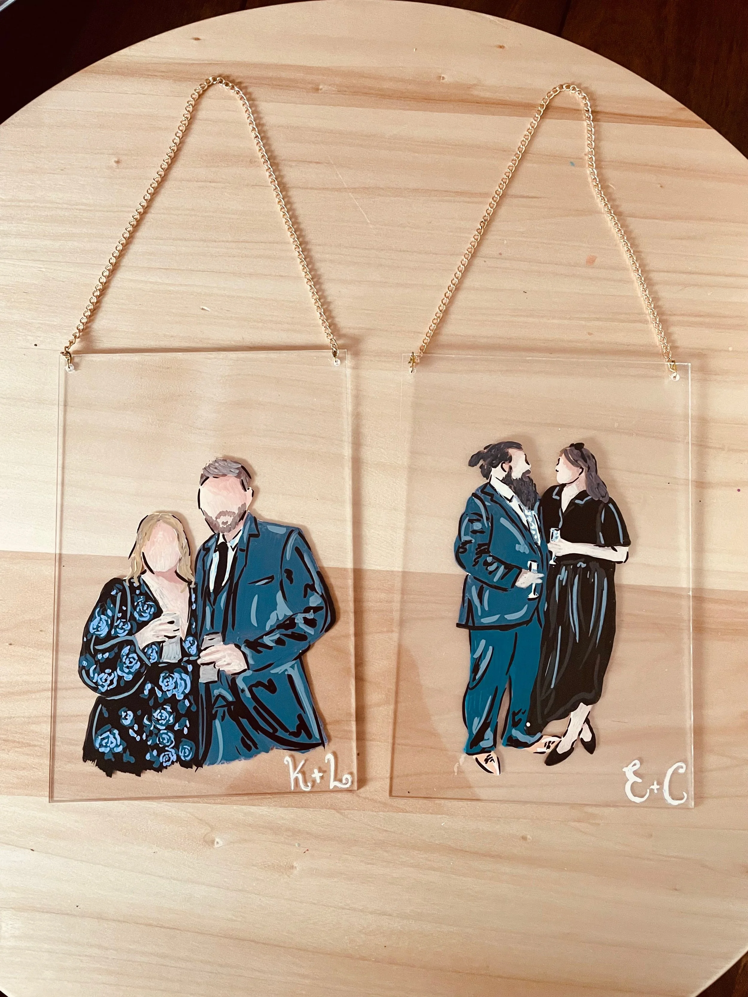 Two sets of acrylic jewelry with couples' illustrations, one labeled 'K + 2' and the other 'E + C', displayed on a wooden surface.
