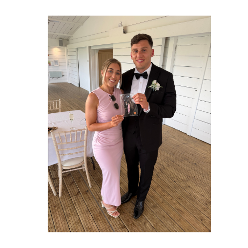 A couple at a wedding, the woman in a pink dress and sunglasses, the man in a tuxedo, holding a guest illustration standing next to a table with gold chairs in a bright white room with wooden floors.