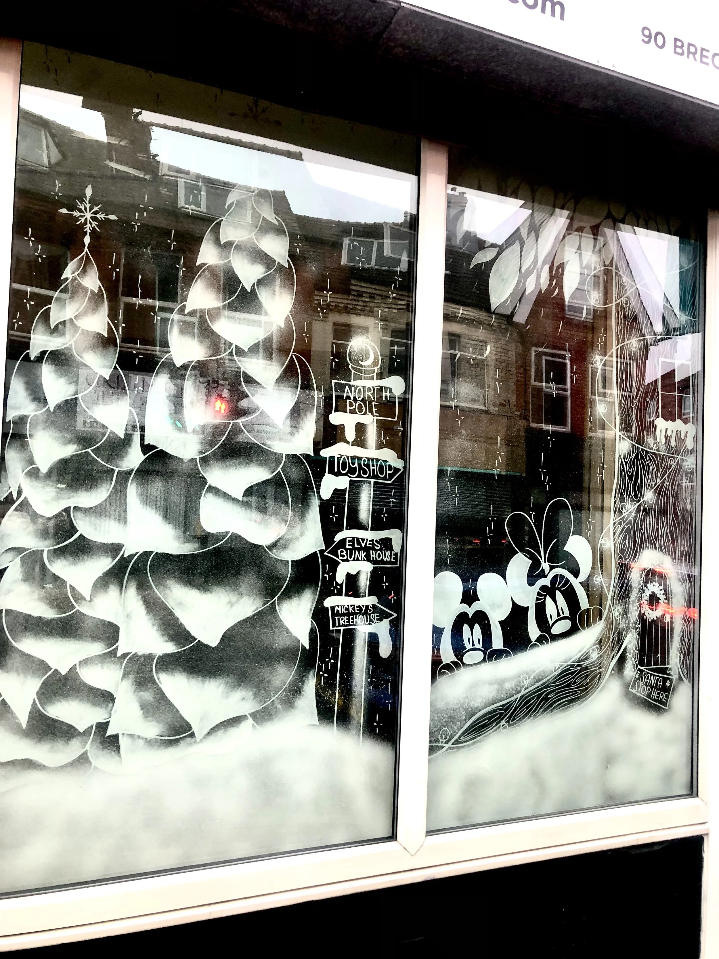 Display window with white Christmas-themed illustrations including pinecones, Santa's sleigh, and Mickey Mouse signs pointing to North Pole, toy shop, elves' bunk house, Mickey's treehouse, and Santa's workshop.