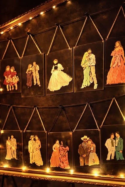 Collection of framed embroidered or painted portraits of couples in vintage or country-style clothing on a dark wall with string lights above and below.