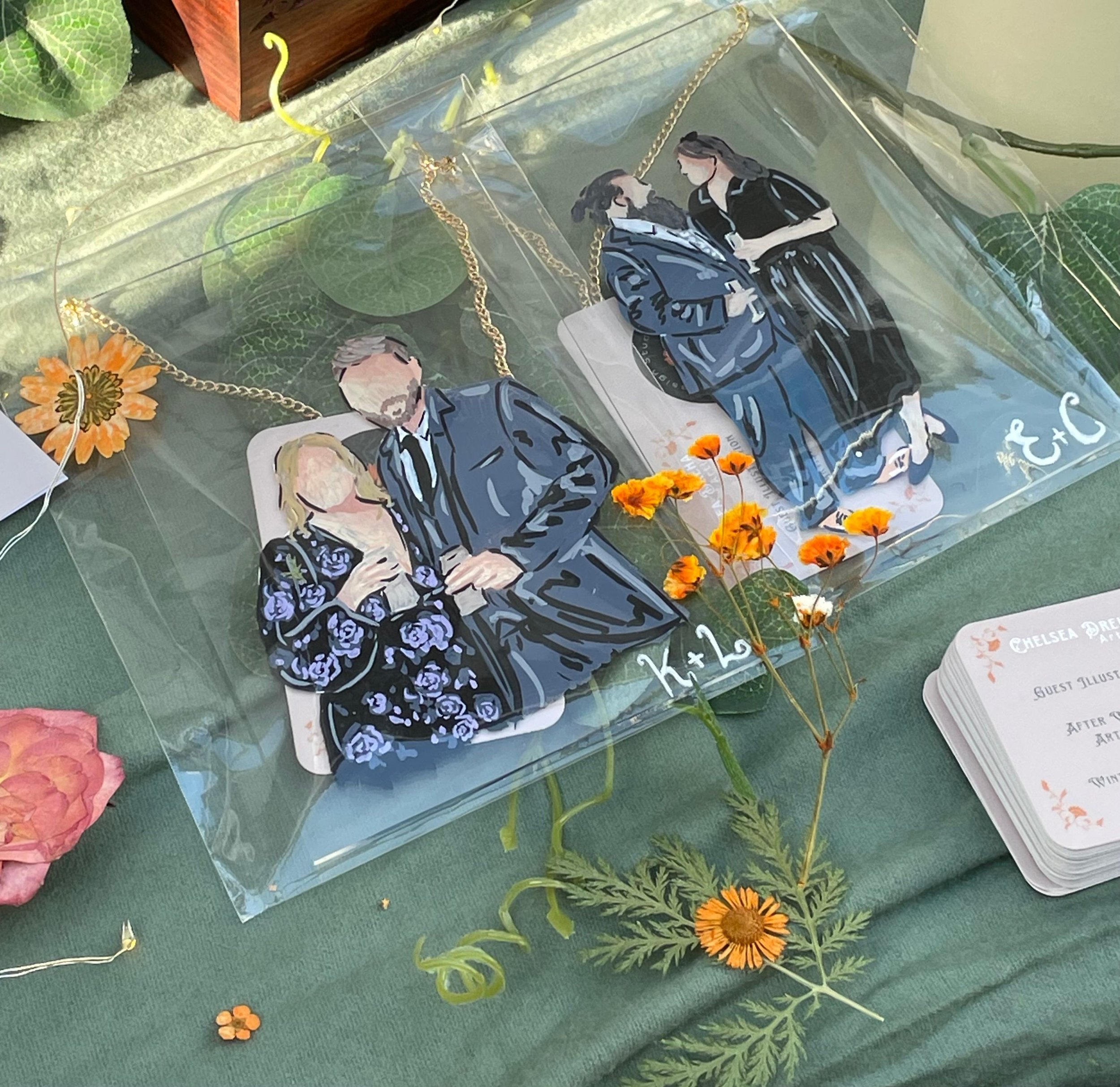 Three illustrated jewelry items featuring couples, displayed on a green surface with flowers, leaves, and a card nearby.