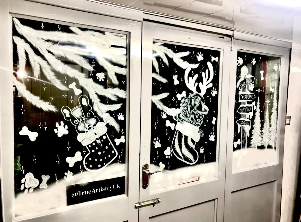 Decorated windows with white holiday-themed illustrations, including a dog in a stocking, a lion with antlers, and directional signs, surrounded by pine branches and paw prints.