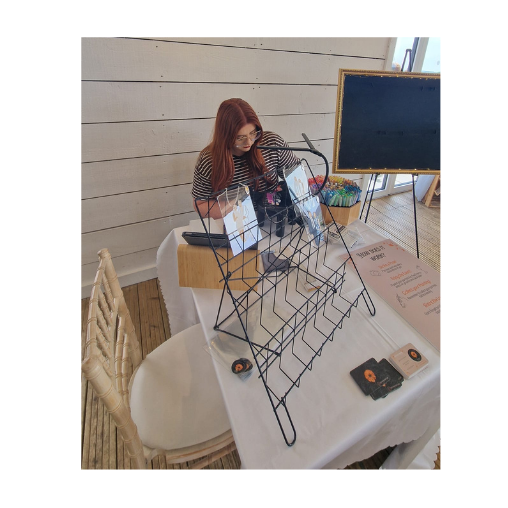 A young woman with red hair and glasses is standing at a table with a white tablecloth, looking down at a display stand. Live wedding Illustrator