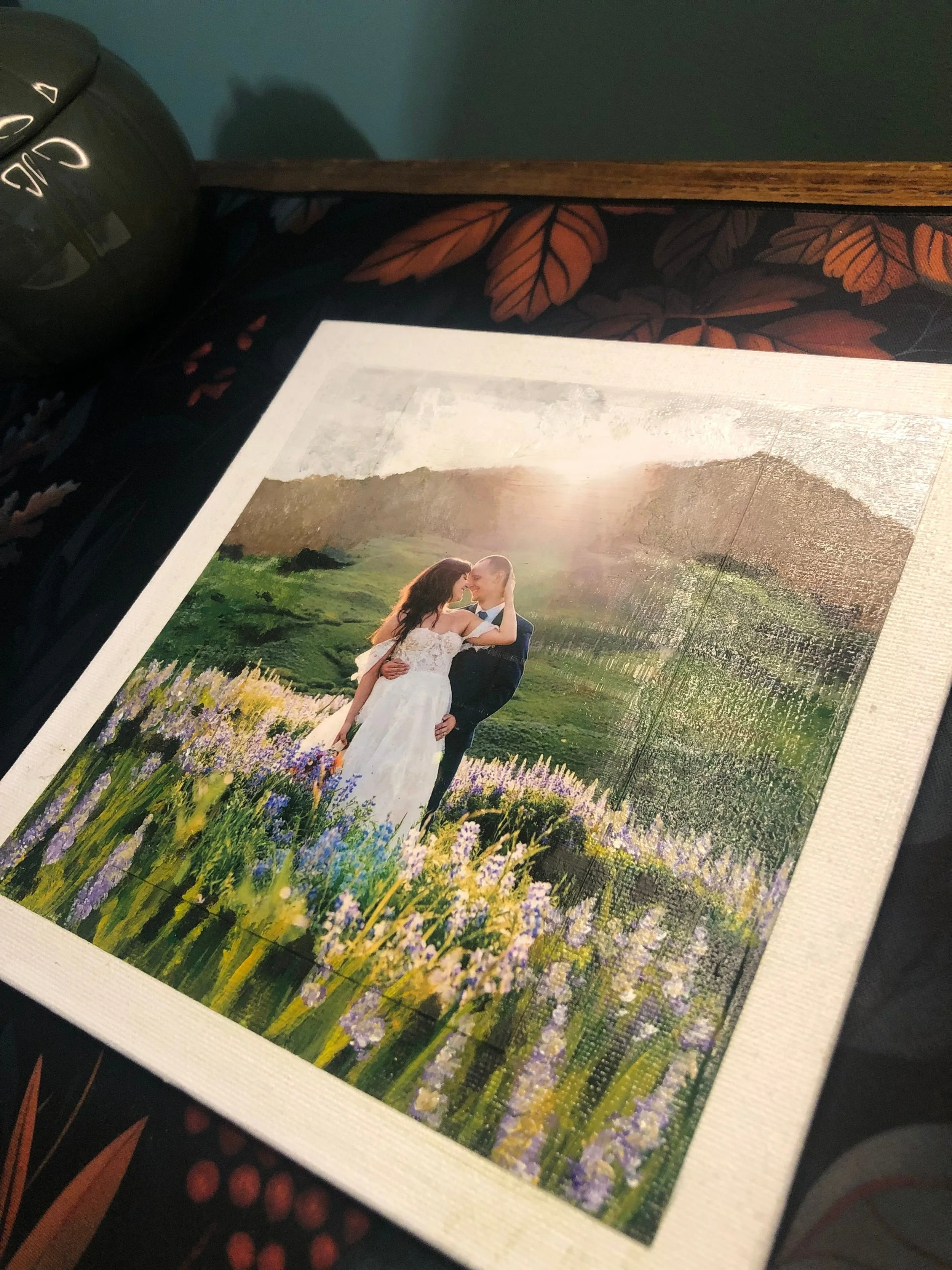 Printed photograph of a bride and groom embracing in a field of wildflowers with a mountain and sunset in the background.
