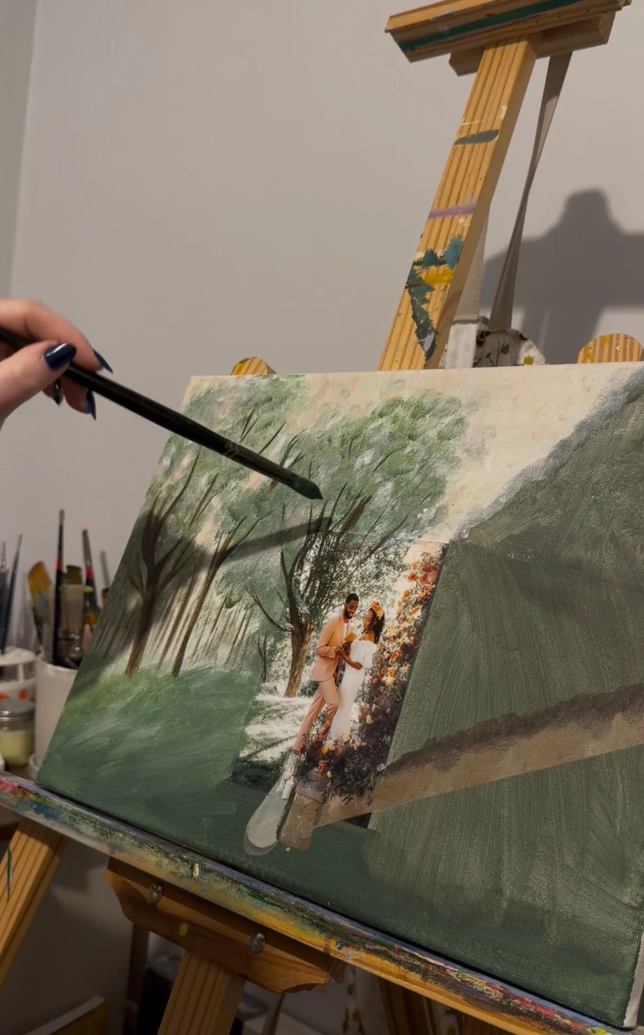 An artist paints a landscape scene on a canvas, which features a couple in formal attire standing outdoors among trees and greenery. There are art supplies and brushes in the background.