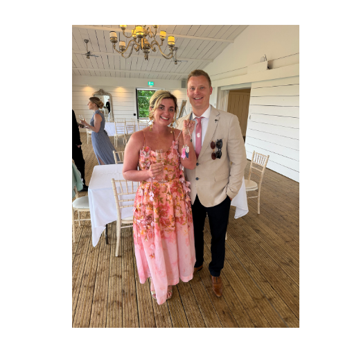 Guests holding their live wedding illustration portrait. The woman is wearing a pink floral dress, and the man is dressed in a white blazer, pink tie, and dark pants, with sunglasses hanging from his jacket.