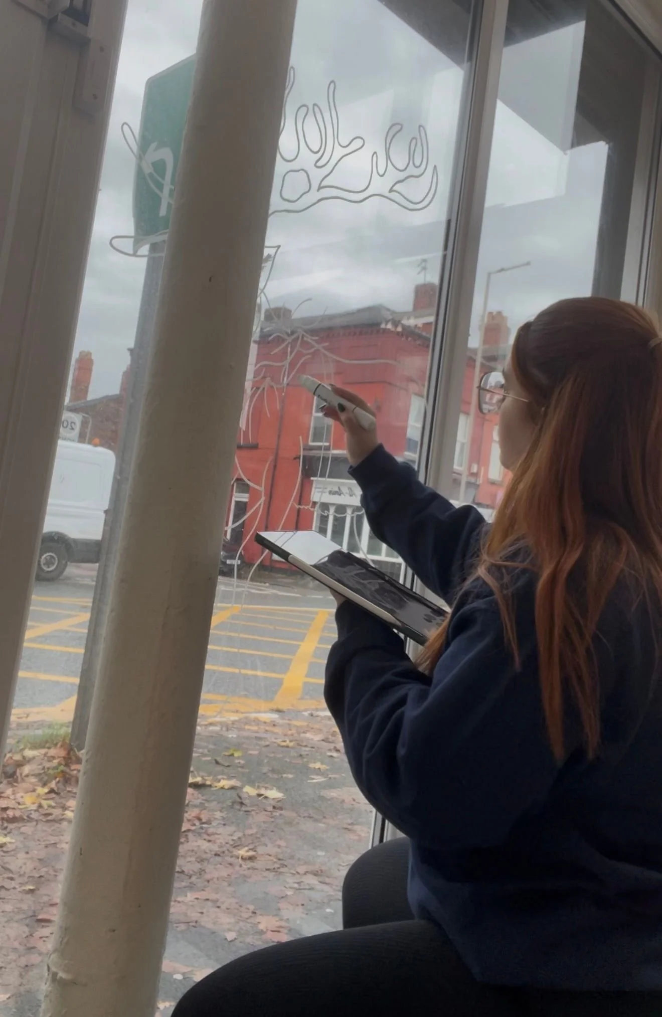 A woman with red hair and glasses drawing a snowflake on a window with a white marker, holding a tablet in her left hand, outside view of a red brick building and parked vehicles.