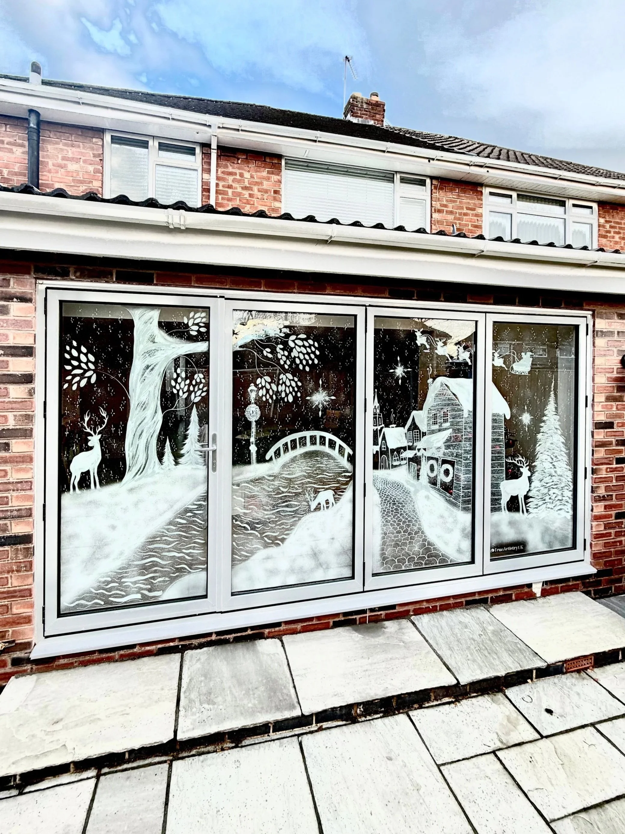 Window decorated with white winter scene featuring a large tree with leafless branches, a deer, a bridge, a river, horses, houses, Christmas trees, and stars, with snow-covered ground.