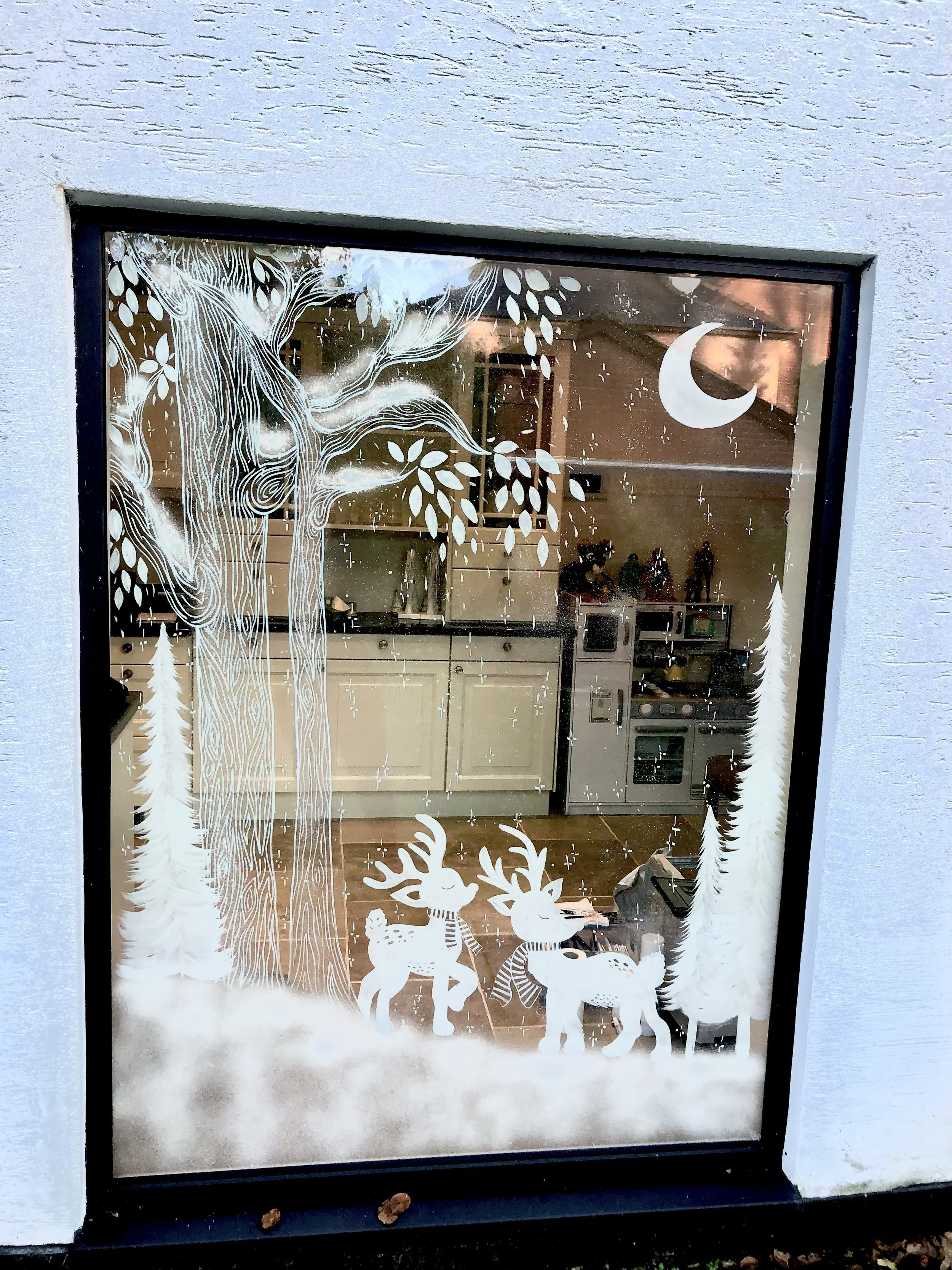 Winter-themed window decoration featuring a large tree, two pine trees, a crescent moon, and two reindeer, all depicted in white on the glass.