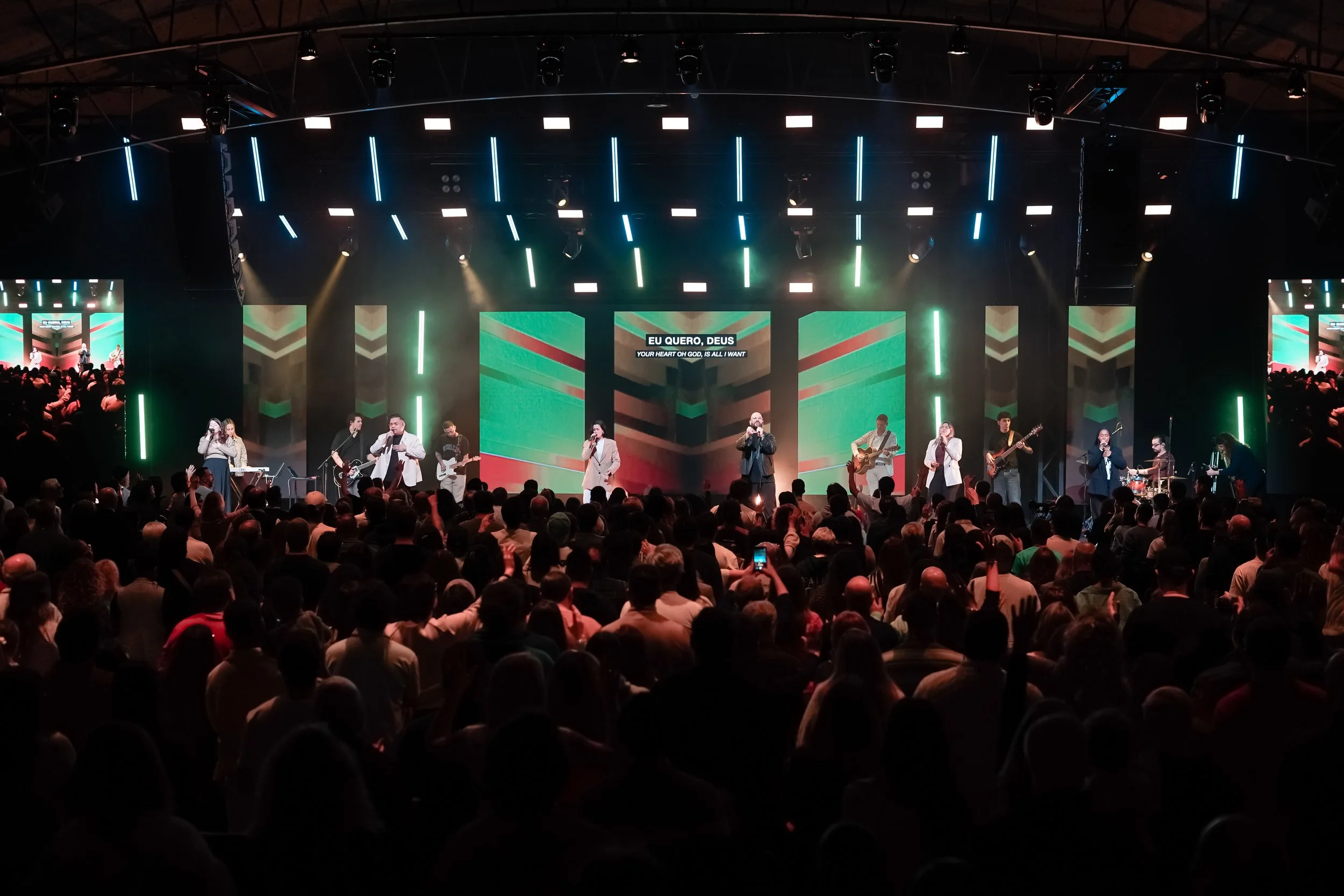 Auditório interior para concertos e eventos, com palco iluminado, LED wall, ecrãs digitais e sistema de som profissional, com músicos em atuação e público presente.