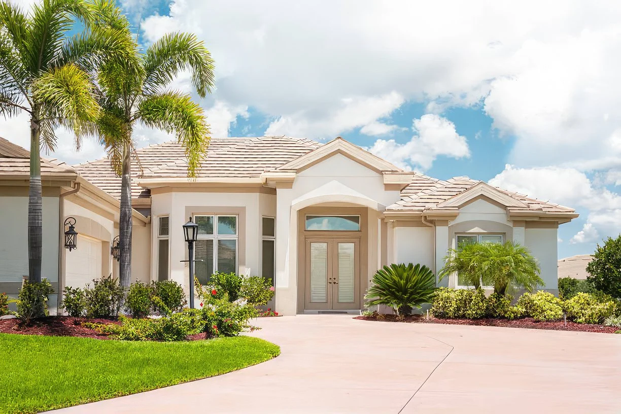 A modern, white house with a tiled roof, front yard with green grass, palm trees, and landscaped bushes, under a partly cloudy sky.