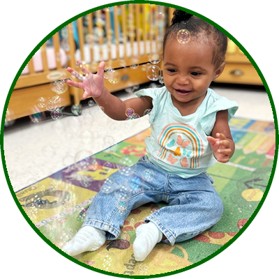 Young girl sitting on a colorful play mat, reaching for bubbles, smiling, with a wooden crib in the background.