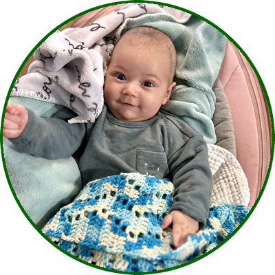 A smiling baby with light skin and short hair, sitting in a cushioned chair and wrapped in a colorful knitted blanket.