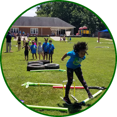 Children participating in outdoor activities at a park, with a girl riding a skateboard in the foreground and other kids waiting in line in the background.