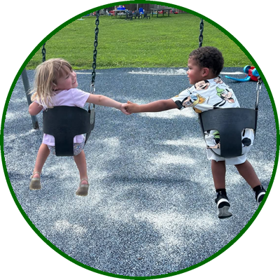 Two young children, a girl and a boy, are holding hands while sitting on swings at a park playground, with green grass and picnic tables in the background.