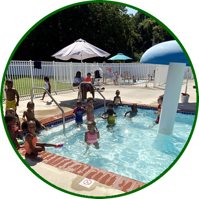 Children swimming and playing in a small outdoor pool with umbrellas and a water slide.