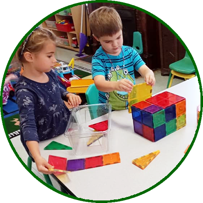 Two children, a girl and a boy, playing with colorful translucent building blocks at a white table in a classroom or playroom.