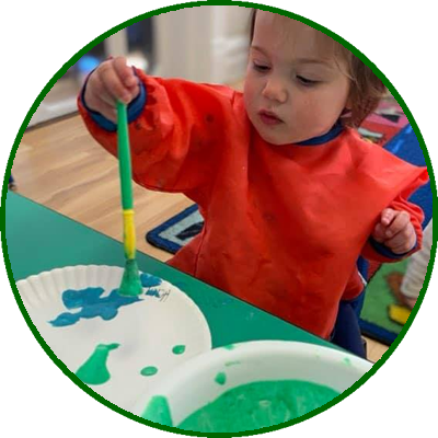 A young child in a red smock is using a paintbrush to paint blue on a paper plate, with containers of green paint nearby.