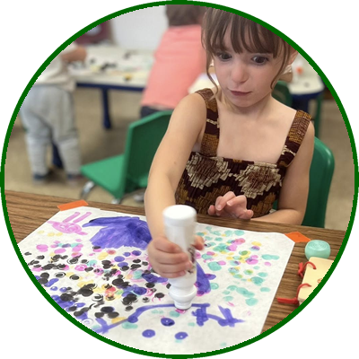 Young girl creating colorful artwork with markers and paint on paper in a classroom.