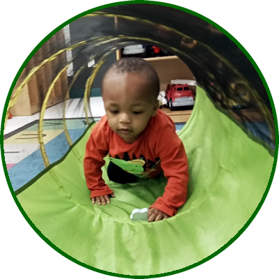 Child crawling through a green play tunnel with a wooden floor, toy cars, and furniture in the background.