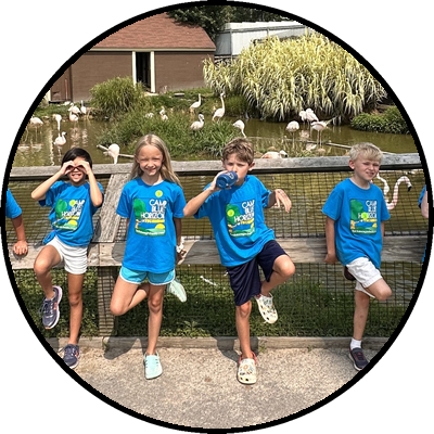 Four children in blue T-shirts, posing playfully in front of a pond with swans, with greenery and a building in the background.