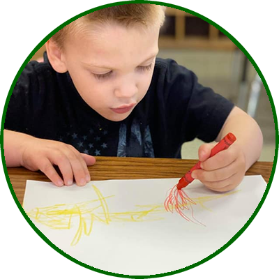 A young boy drawing on a piece of paper with a red crayon.