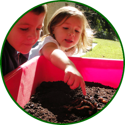 Two children, a boy and a girl, examining and touching soil in a pink gardening container outdoors on a sunny day.