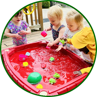 Three young children playing with water and toys in a red plastic sandbox outdoors.