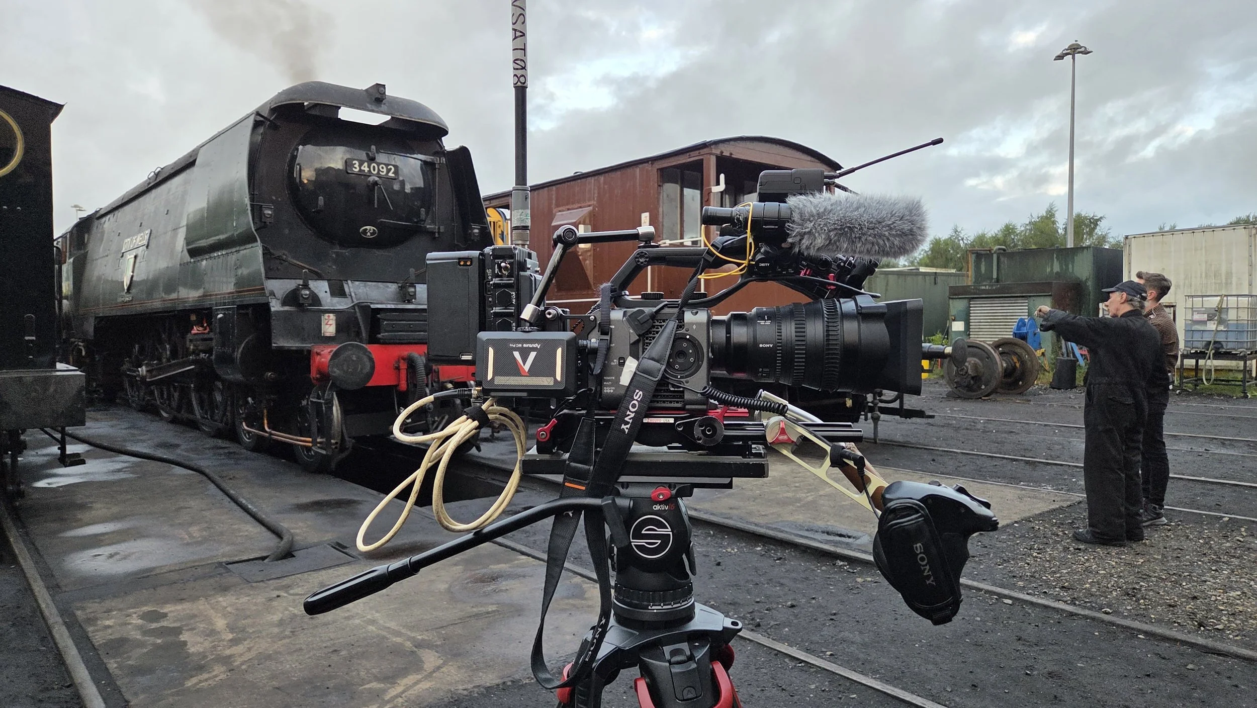 Film crew filming on railroad tracks with vintage steam locomotive and wooden train cars, camera equipment in foreground.