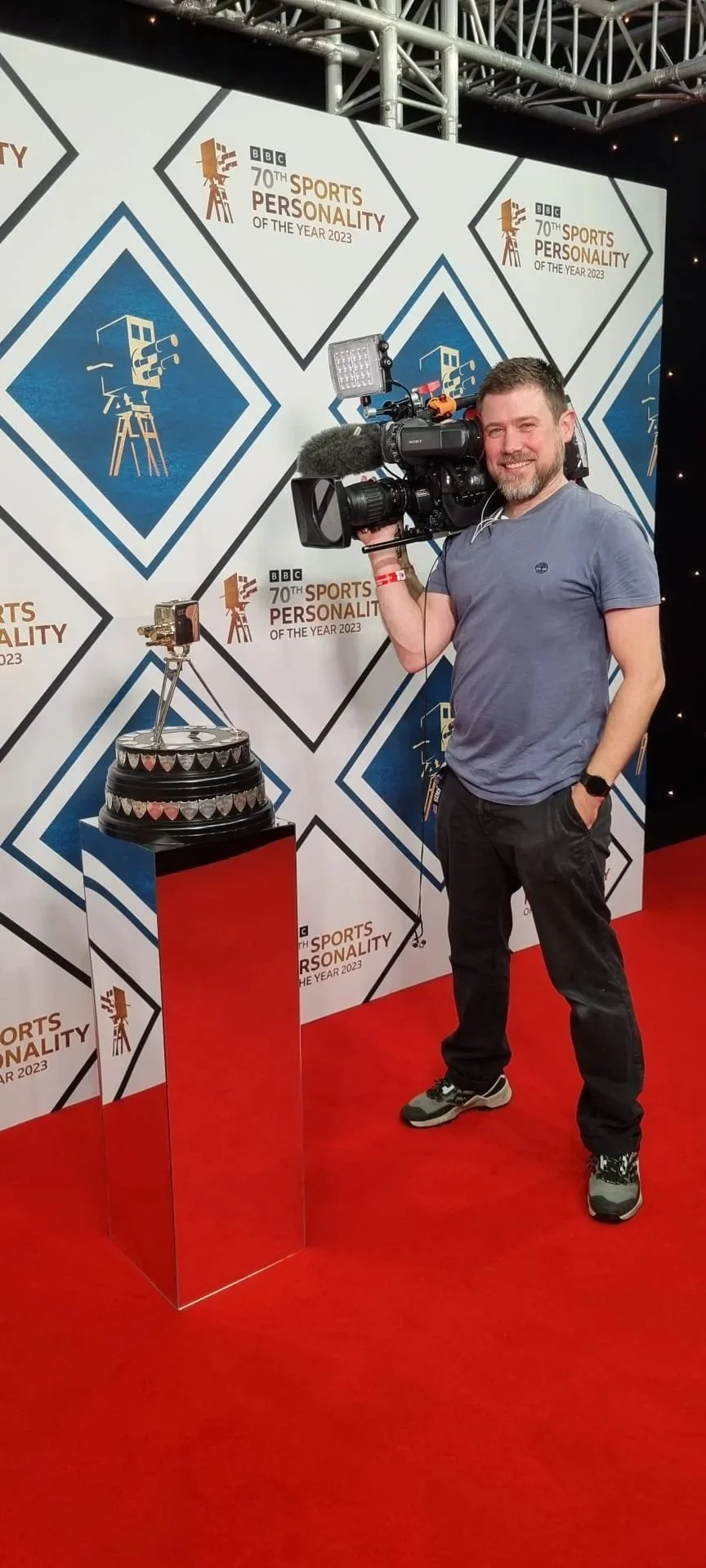 A man standing on a red carpet holding a professional video camera at an event with a backdrop reading "BBC 70th Sports Personality of the Year 2023." There is a trophy on a red podium beside him.