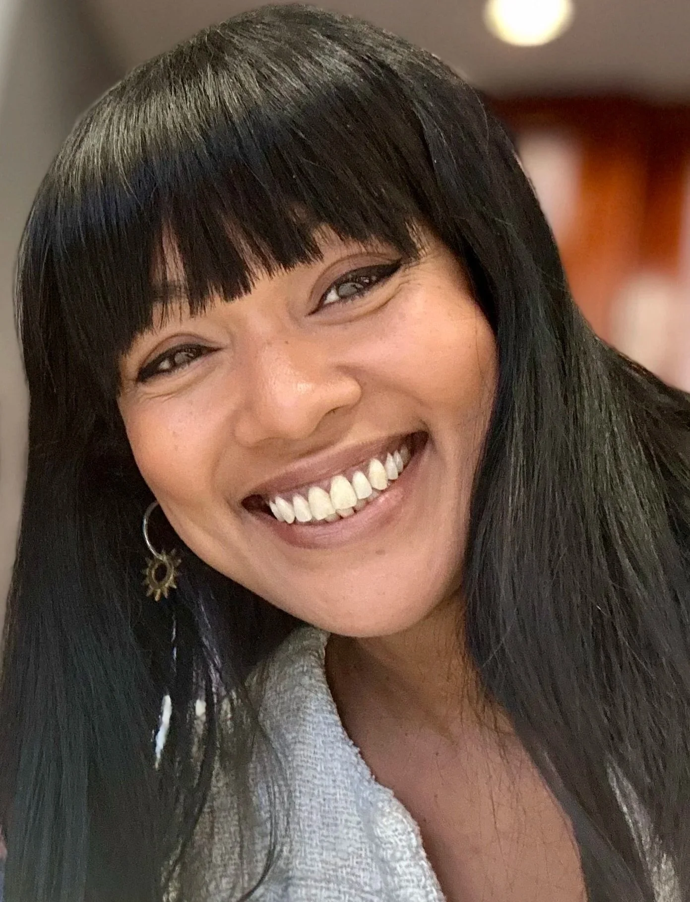 Close-up of a woman smiling with black hair and bangs, wearing earrings and a light-colored top.