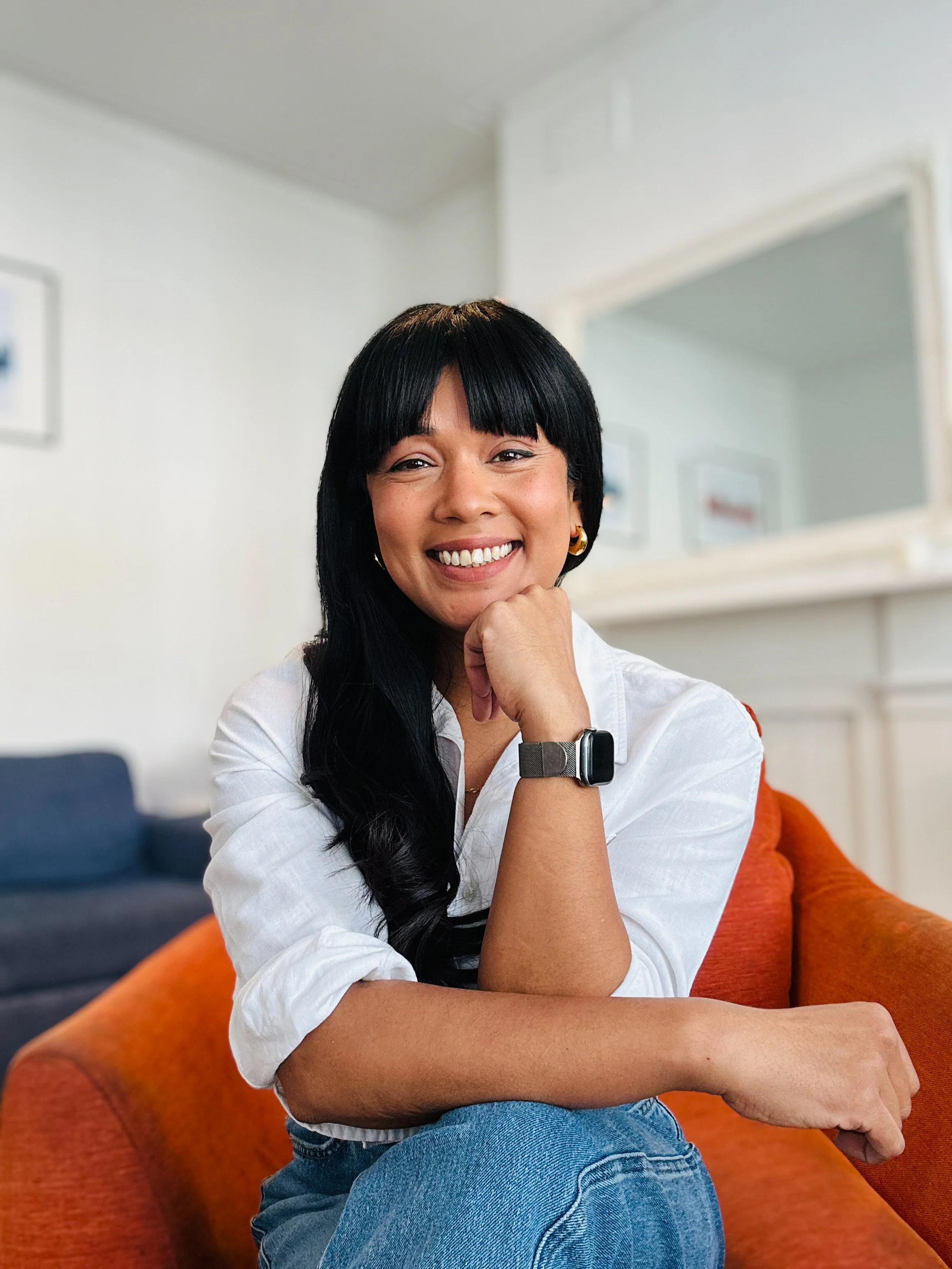 Shu Beckwith, counsellor and psychotherapist in South West London, smiling with her head resting on her right hand in a warm inviting living room with framed pictures on the wall and a mirror.