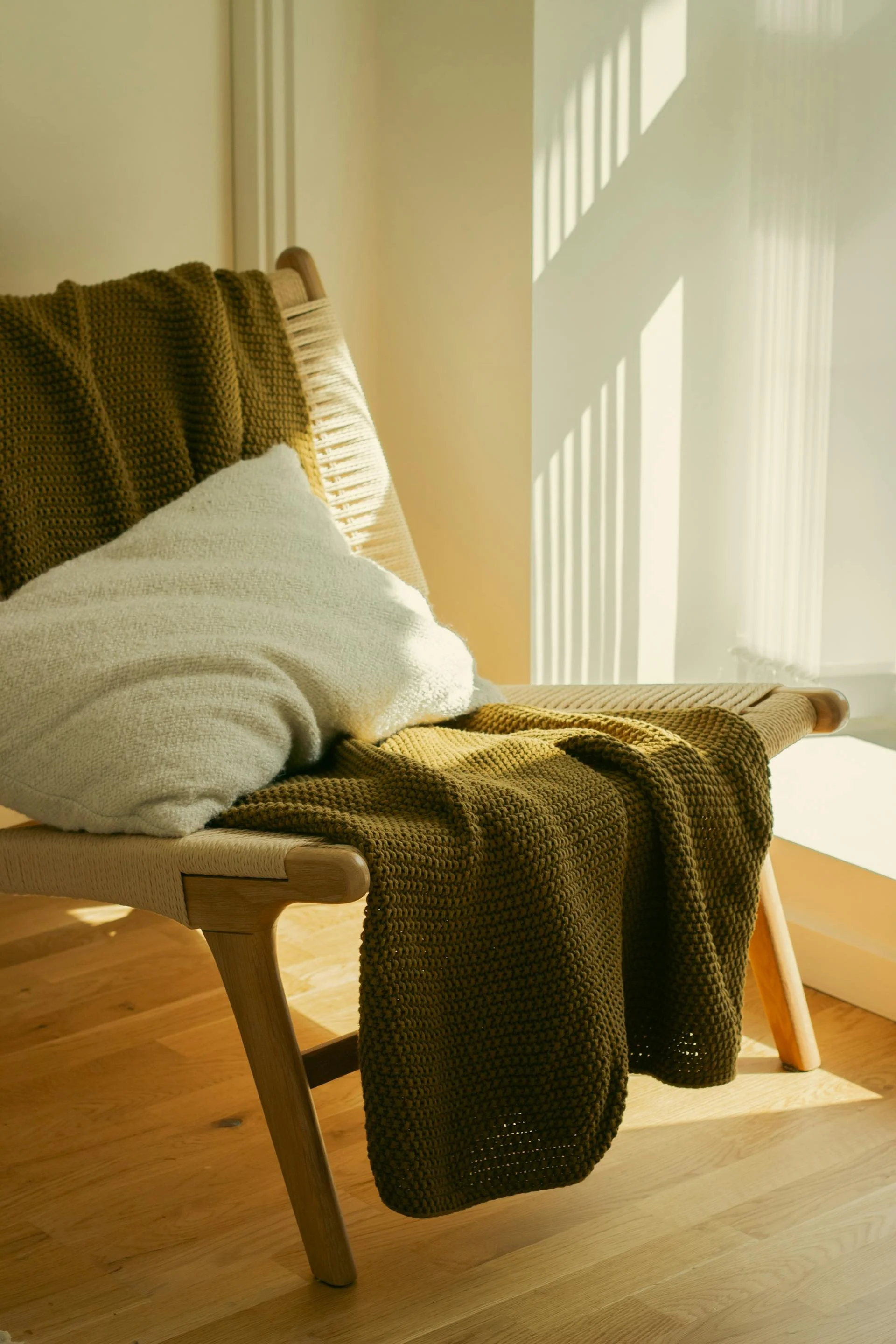 Sunlight streaming through window blinds onto a wooden bench with a draped green knitted blanket and two pillows, one white and one with yellow stripes, in a cozy corner of a room.