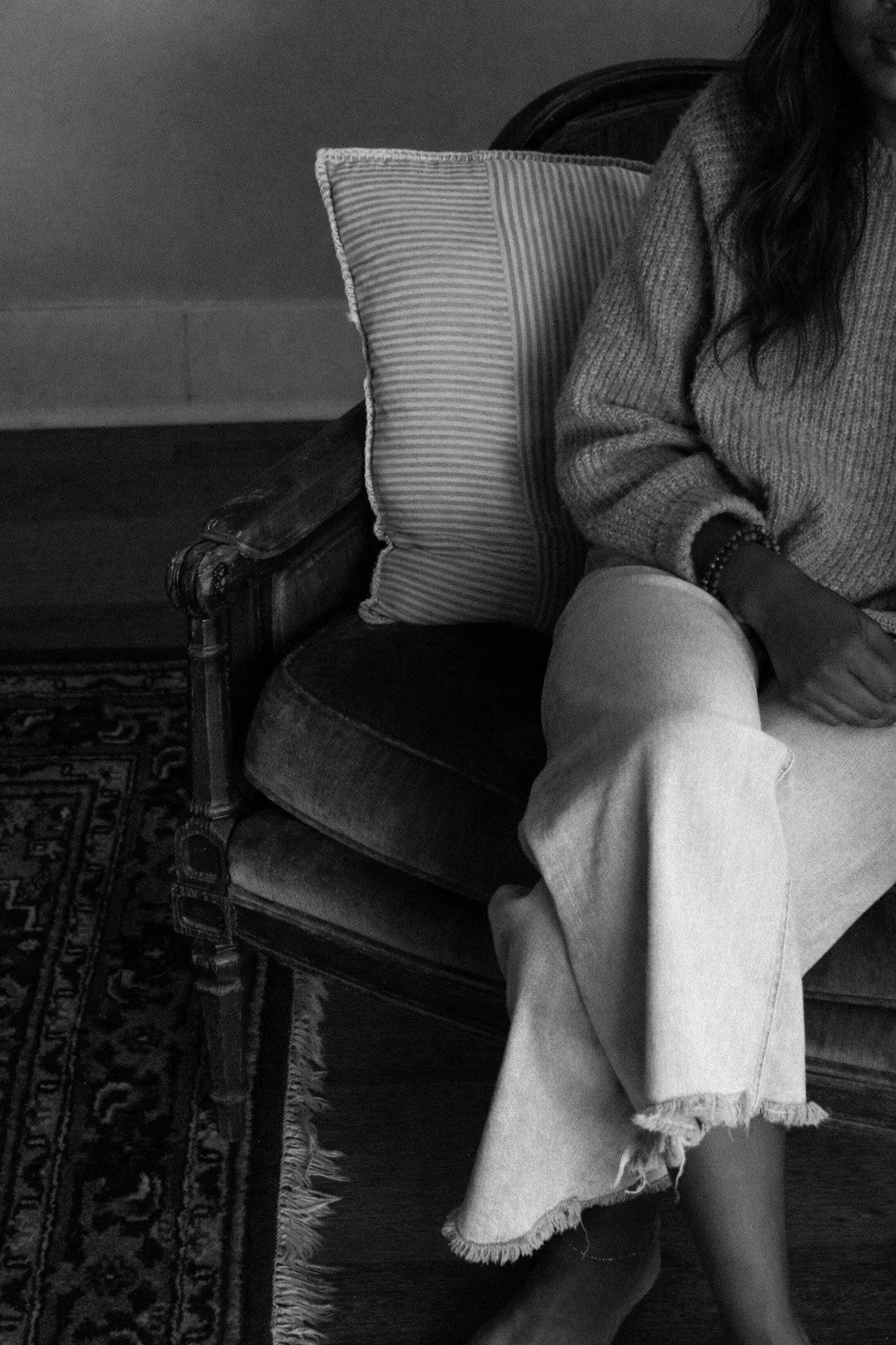 Partial view of a woman sitting on a vintage chair with a striped pillow, wearing a knit sweater and frayed hem jeans, with bracelets on her wrist.